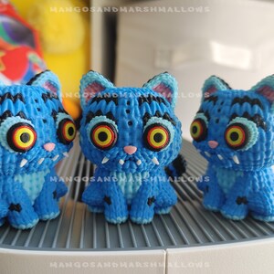 May include: Three blue cat figurines with large, yellow and red eyes, and black stripes. The handmade cats have white fangs, pink noses, and black and blue striped tails. The figurines are sitting on a white surface.
