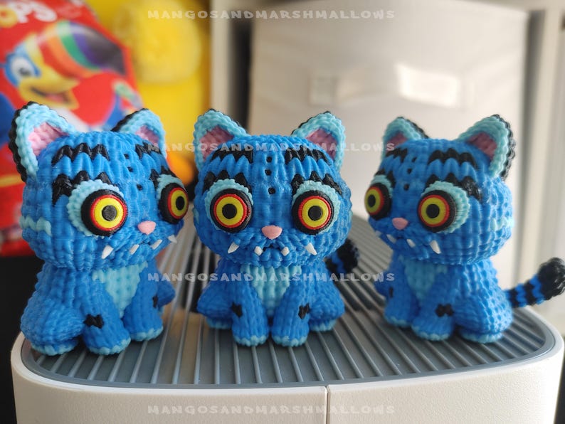 May include: Three blue cat figurines with large, yellow and red eyes, and black stripes. The handmade cats have white fangs, pink noses, and black and blue striped tails. The figurines are sitting on a white surface.