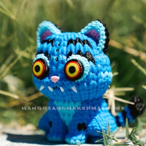 May include: A handmade blue crochet cat with black stripes and yellow and red eyes. The cat has a pink nose and white fangs. The text "MANGOSANDMARSHMALLOWS" is visible at the bottom.