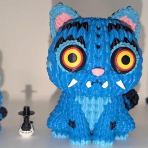 May include: Three whimsical figurines: two blue cat-like creatures with large yellow and red eyes, and a small black and white figure with a hat. The cats have pink ears and white fangs. The figurines are displayed on a white shelf.