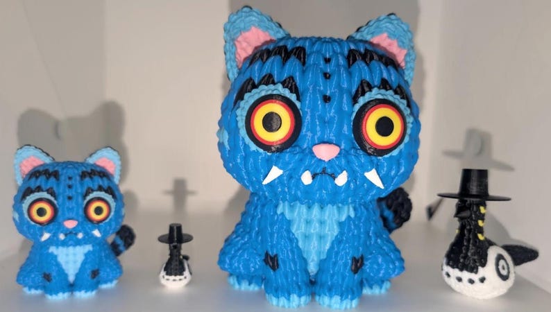 May include: Three whimsical figurines: two blue cat-like creatures with large yellow and red eyes, and a small black and white figure with a hat. The cats have pink ears and white fangs. The figurines are displayed on a white shelf.