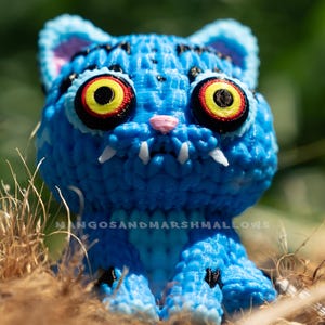May include: A blue cat figurine with large yellow, red, and black eyes, white fangs, and a pink nose. The figurine is textured and has black markings. The text "MANGOSANDMARSHMALLOWS" is visible. The cat is sitting in dry grass.