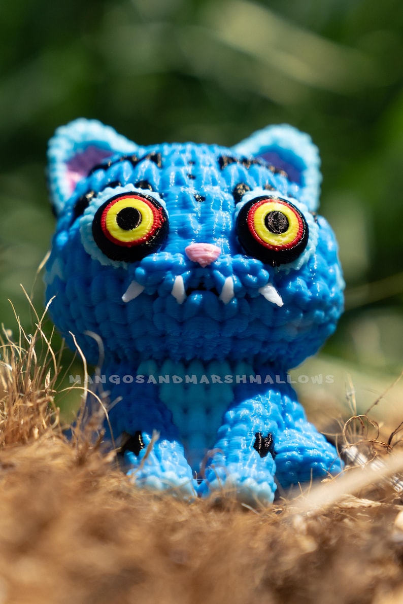 May include: A blue cat figurine with large yellow, red, and black eyes, white fangs, and a pink nose. The figurine is textured and has black markings. The text "MANGOSANDMARSHMALLOWS" is visible. The cat is sitting in dry grass.