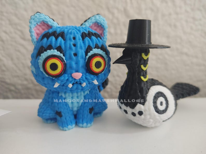 May include: A blue cat figurine with black markings and large yellow eyes, next to a black and white bird figurine wearing a black top hat. The cat has a pink nose and ears. The bird has yellow accents.