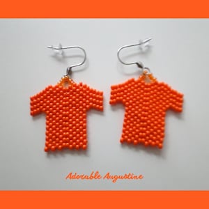 May include: A pair of earrings shaped like t-shirts, crafted from small, vibrant orange beads. Each earring features a silver-toned metal hook with a clear plastic backing. The text "Adorable Augustine" is printed in orange below the earrings.