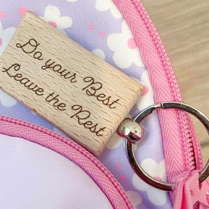 May include: A rectangular wooden keychain with the engraved text "Do your Best Leave the Rest." The keychain has a silver-colored ring and a small silver-colored bell. The background is a pink zipper and a floral pattern.