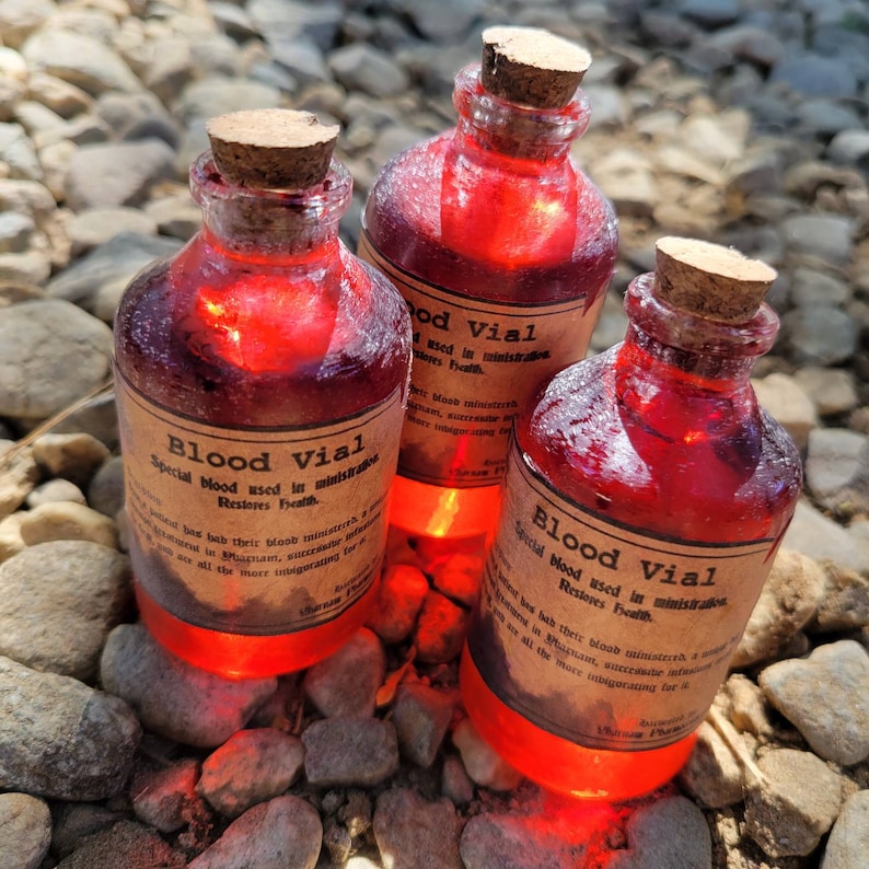 May include: Three glass vials filled with a glowing red liquid, each sealed with a cork stopper. The vials have labels that read "Blood Vial" with additional text. The vials are resting on a bed of small, gray stones.