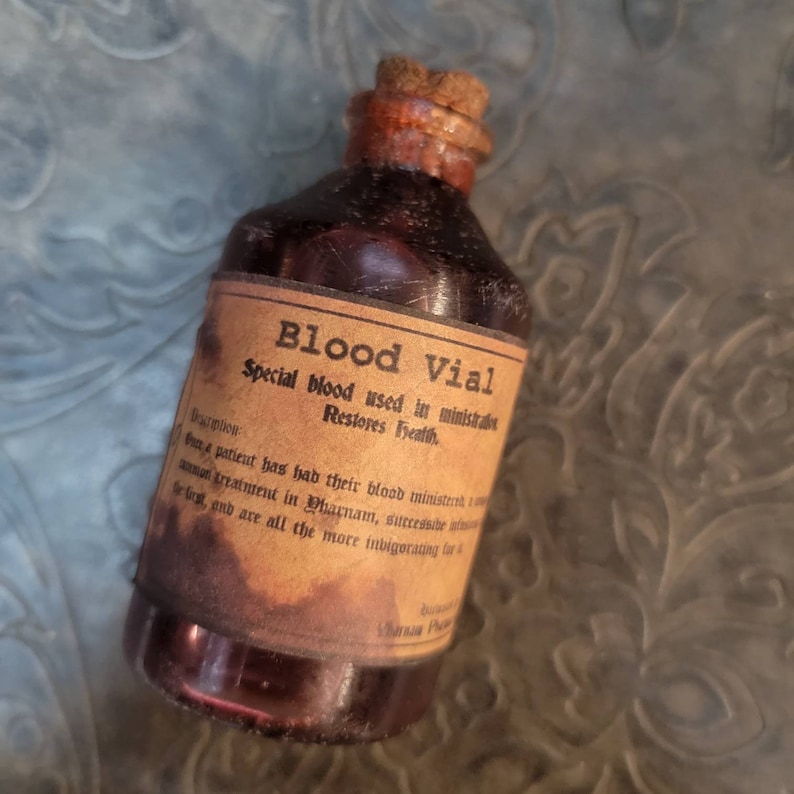 May include: A vintage-style glass bottle labeled "Blood Vial" with a cork stopper. The label reads "Special blood used in ministrations, Restores Health." The bottle has a brown hue and appears aged, with text that reads "Description" and additional text below.