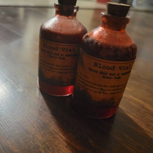May include: Two antique-style glass bottles filled with a red liquid, labeled "Blood Vial." The labels are aged with text that reads "Special blood used in... Restores Health." Each bottle has a cork stopper.
