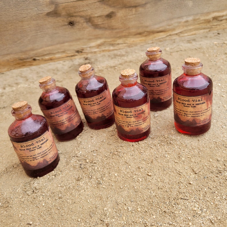 May include: Six small, round glass bottles filled with a deep red liquid, each sealed with a cork stopper. The bottles have aged-looking labels that read "Blood Vial". The bottles are arranged on a sandy surface.