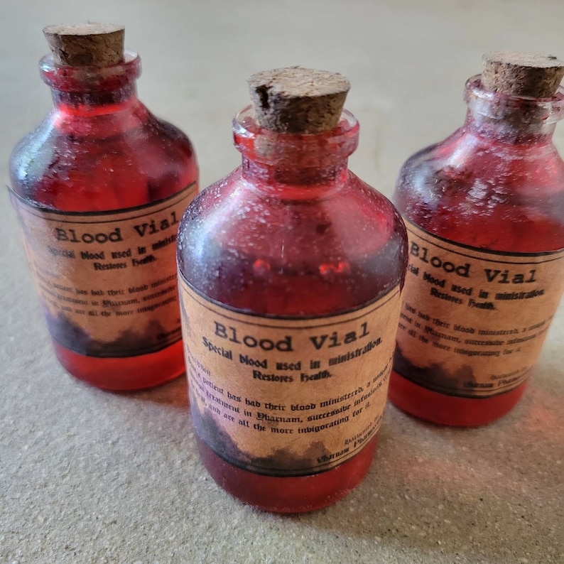 May include: Three small, red glass bottles with cork stoppers. Each bottle has a label that reads "Blood Vial" and includes additional text. The bottles are filled with a red liquid, and the labels have a vintage appearance.