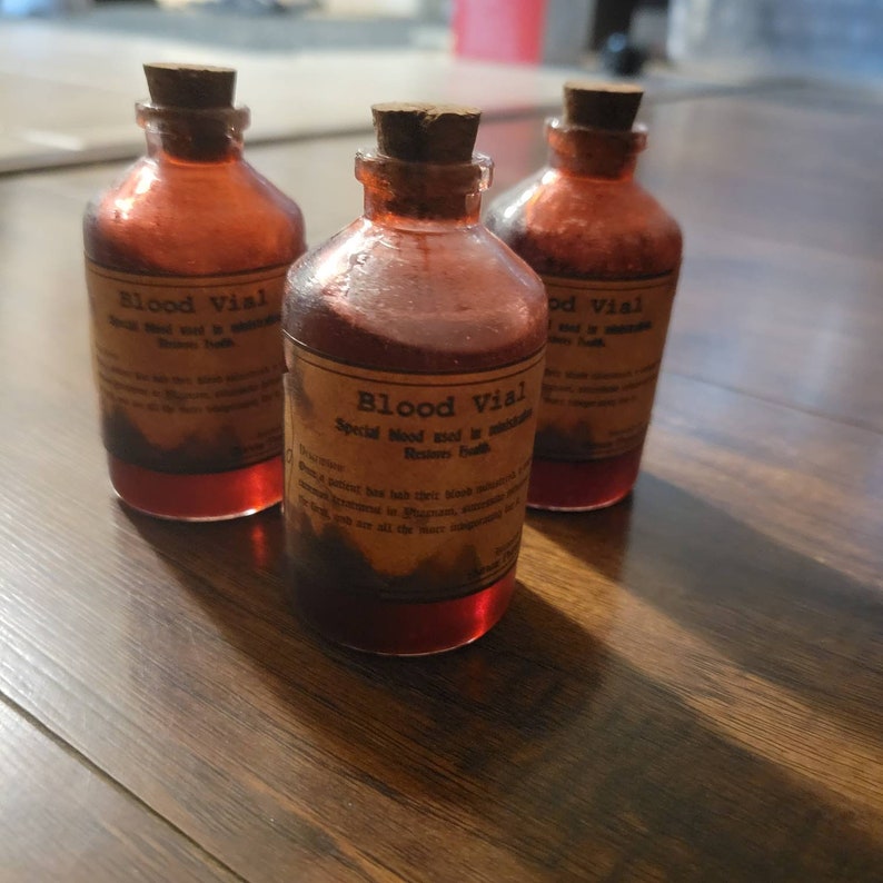 May include: Three glass bottles with cork stoppers, labeled "Blood Vial." The bottles are filled with a red liquid, and the labels have a vintage, aged appearance. The bottles are arranged on a wooden surface.