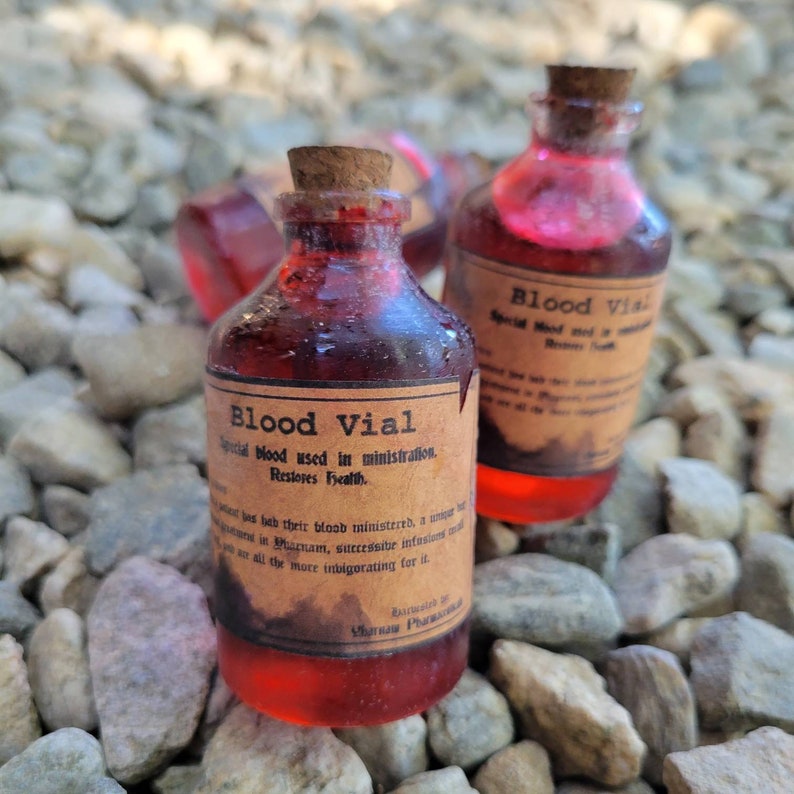 May include: Small glass bottles filled with a red liquid, labeled "Blood Vial" with cork stoppers. The labels have aged, vintage-style text. The bottles are arranged on a bed of small, light-colored stones.
