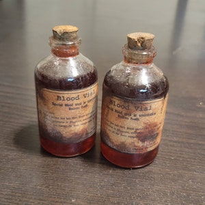 May include: Two small glass vials filled with a dark red liquid, each sealed with a cork stopper. The vials have aged, distressed labels that read "Blood Vial" and "Restores Health."