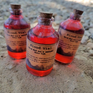 May include: Three glass vials filled with a vibrant red liquid, each sealed with a cork stopper. The vials have labels that read "Blood Vial" with additional text. The bottles are arranged on a sandy surface, creating a mystical or apothecary theme.