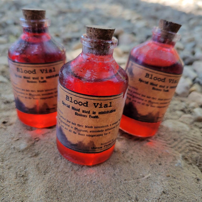 May include: Three glass vials filled with a vibrant red liquid, each sealed with a cork stopper. The vials have labels that read "Blood Vial" with additional text. The bottles are arranged on a sandy surface, creating a mystical or apothecary theme.
