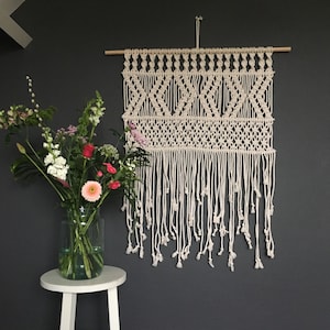 Op de afbeelding: Een witte macramé wandhanger met een geometrisch patroon en franjes. De hanger is gemaakt van katoenen koord en is ongeveer 61 cm breed en 91 cm lang.
