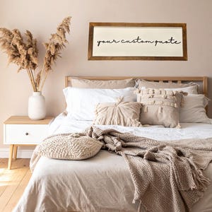 May include: A rectangular wall sign with a dark wood frame and a white background featuring the black cursive text your custom quote. The sign is mounted on a light beige wall above a bed with white and beige bedding, pillows, and a textured throw.