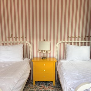 May include: A bedroom scene with two white beds, a yellow nightstand, and a lamp. The beds have white bedding and pillows. The nightstand has three drawers. The wall is striped pink and white. The names "olive" and "evie jo" are on the wall.