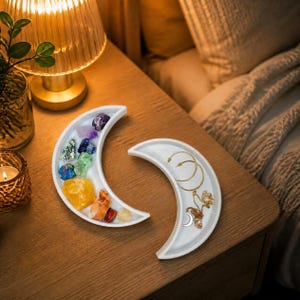 May include: Two white crescent moon-shaped trays. The left tray holds a collection of raw, multi-colored gemstones including purple, blue, green, yellow, and orange stones. The right tray contains gold-toned jewelry, specifically two hoop earrings and a delicate chain necklace with a small crescent moon pendant.