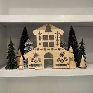 May include: A wooden Christmas village scene with a cutout house, trees, and a bell on the roof. The house has a window and a door, and the trees are decorated with snow.