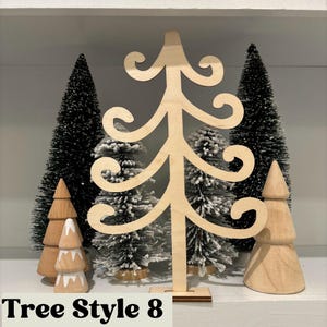 May include: A wooden Christmas tree cutout with a whimsical design. The tree is standing on a shelf with other smaller wooden trees and artificial pine trees.