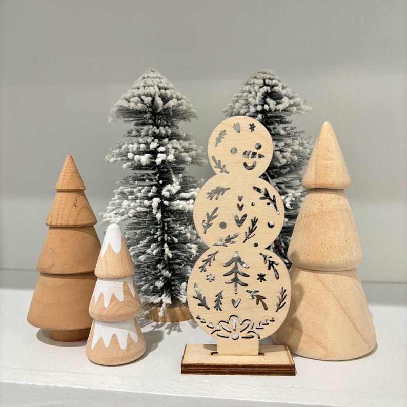 May include: A wooden snowman ornament with a white painted face and a silver glitter accent. The snowman is surrounded by three small wooden Christmas trees, two with white paint and one with a natural wood finish.