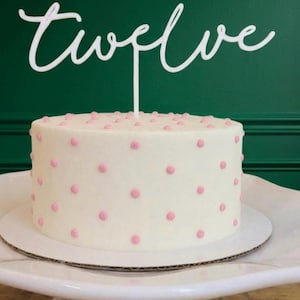 May include: A white cake topper featuring the word twelve in a cursive script font, positioned atop a round white cake decorated with small pink polka dots. The topper has a single thin stem that is inserted into the center of the cake.
