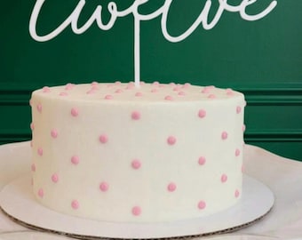 Op de afbeelding: Een witte taarttopper met het woord twelve in een cursief lettertype, geplaatst op een ronde witte taart versierd met kleine roze stippen. De topper heeft een enkele dunne steel die in het midden van de taart is gestoken.