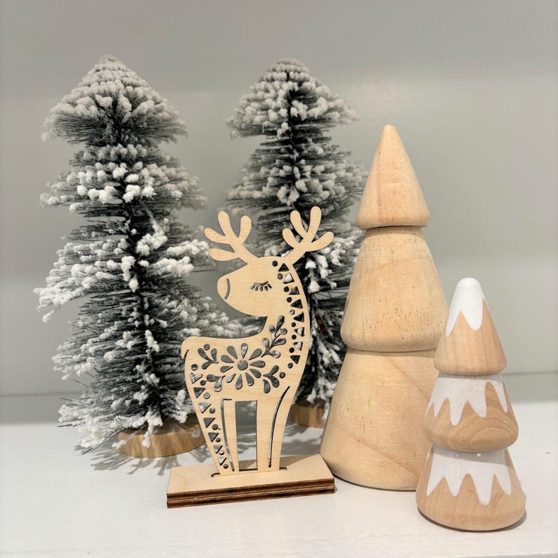 May include: A wooden reindeer figurine with a decorative floral design stands in front of three miniature wooden Christmas trees. The trees are decorated with white snow accents.