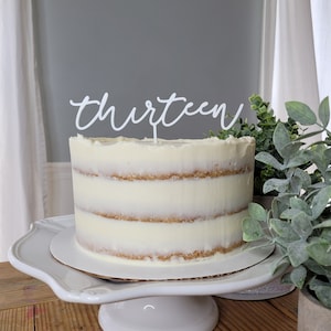 May include: A white cake with white frosting and a white topper that says "thirteen" on a white cake stand. The cake is on a wooden table with a green plant in a pot next to it.