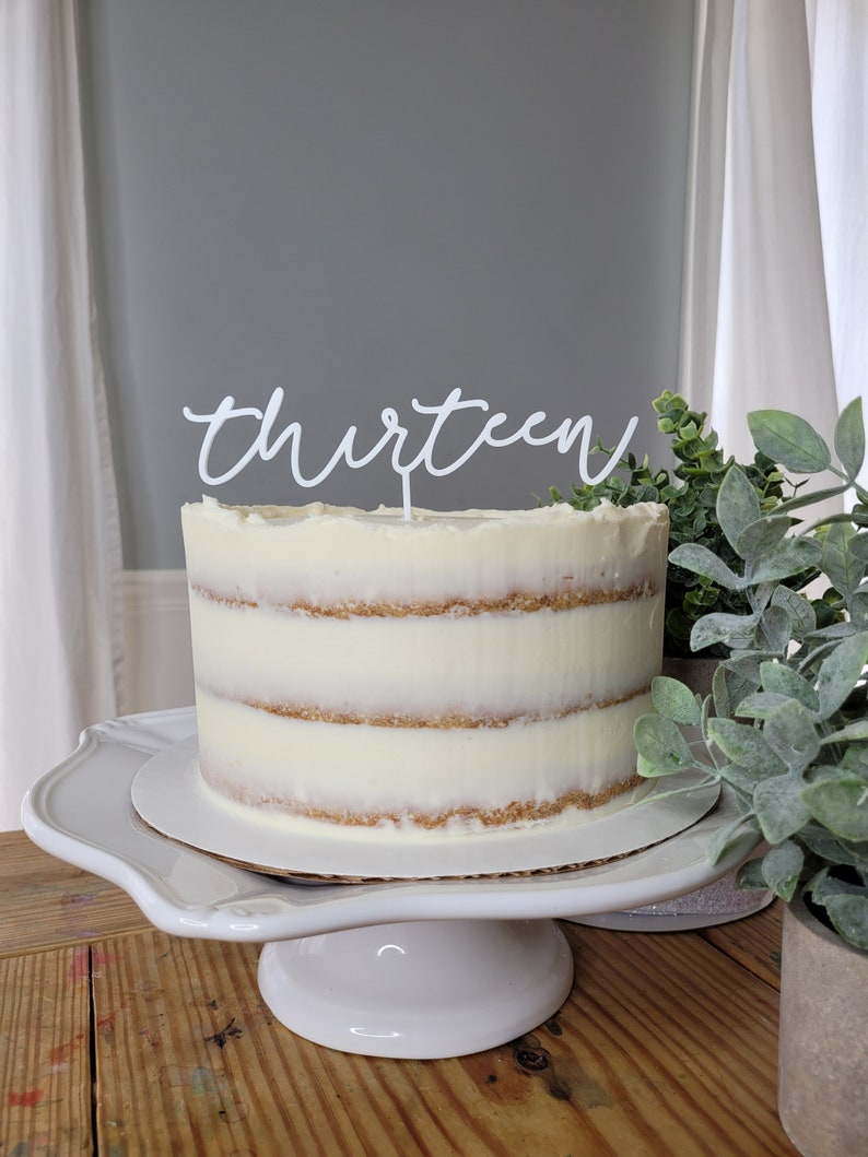 May include: A white cake with white frosting and a white topper that says "thirteen" on a white cake stand. The cake is on a wooden table with a green plant in a pot next to it.