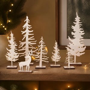 May include: A collection of six light-colored wooden cutouts featuring five evergreen trees of varying heights and one deer silhouette. Each piece is mounted on a small, flat rectangular wooden base, creating a set of freestanding decorative figures.