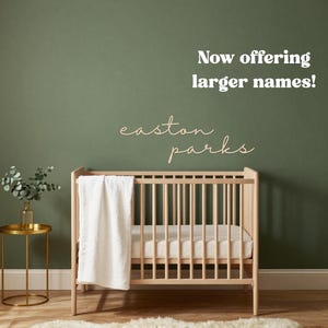 Op de afbeelding: Een lichthouten wieg met een witte deken, geplaatst voor een donkergroene muur met de houten tekst "easton parks" en de witte tekst "Now offering larger names!" erboven, met een gouden bijzettafel en een wit kleed.