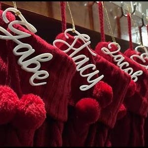 May include: A row of deep red knitted Christmas stockings, each featuring a white, script-style name tag hanging from a gold string. The names visible on the tags include Gabe, Lacy, Jacob, and Ethan, with each stocking accented by two red pom-poms.