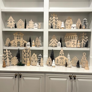 May include: A set of wooden Christmas village decorations featuring houses, trees, and other festive elements. The decorations are painted in a white and gray color scheme with black accents. The decorations are arranged on three shelves of a white bookcase.