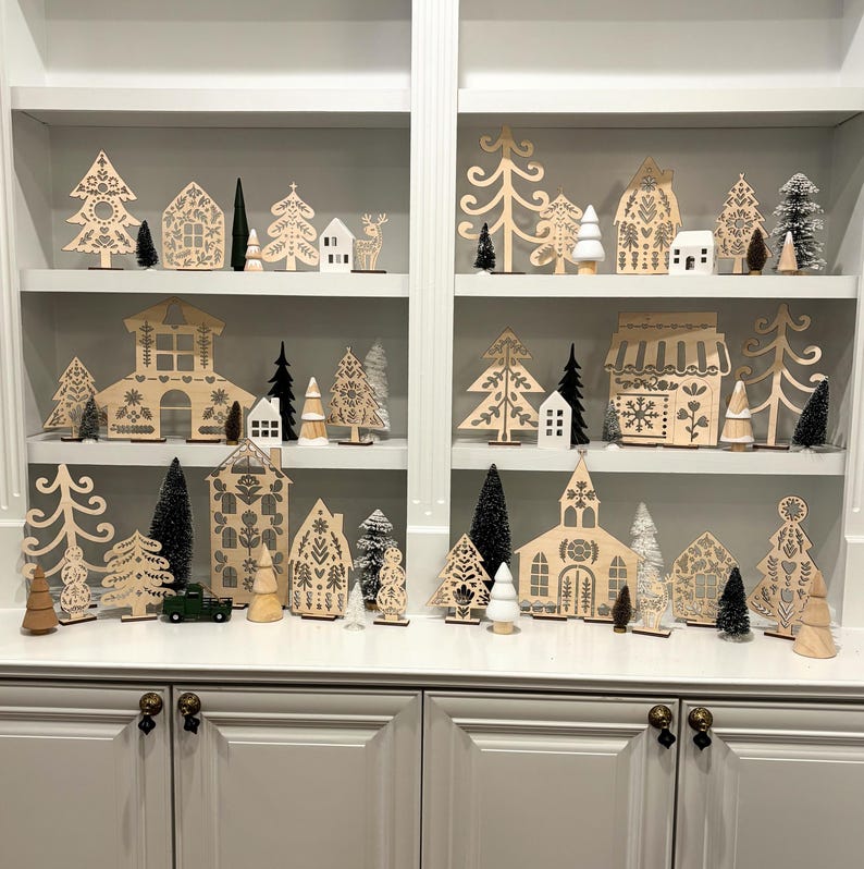 May include: A set of wooden Christmas village decorations featuring houses, trees, and other festive elements. The decorations are painted in a white and gray color scheme with black accents. The decorations are arranged on three shelves of a white bookcase.