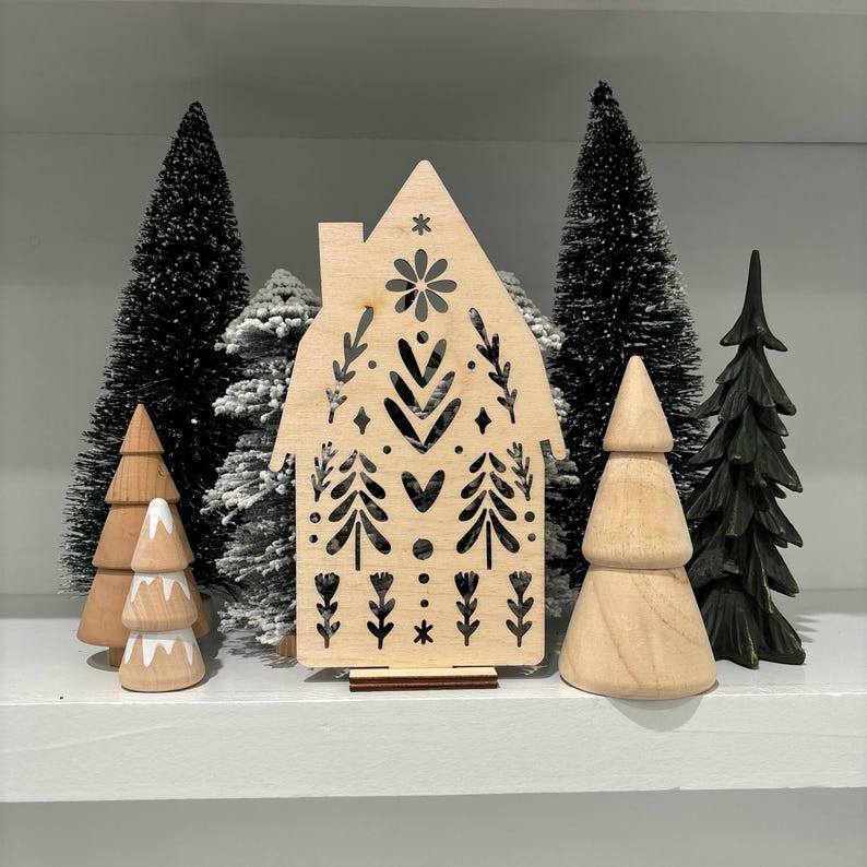 May include: A wooden cutout of a house with a Scandinavian-inspired design. The house is decorated with black and white geometric patterns, including hearts, stars, and trees. The cutout is standing on a shelf with several small artificial pine trees.