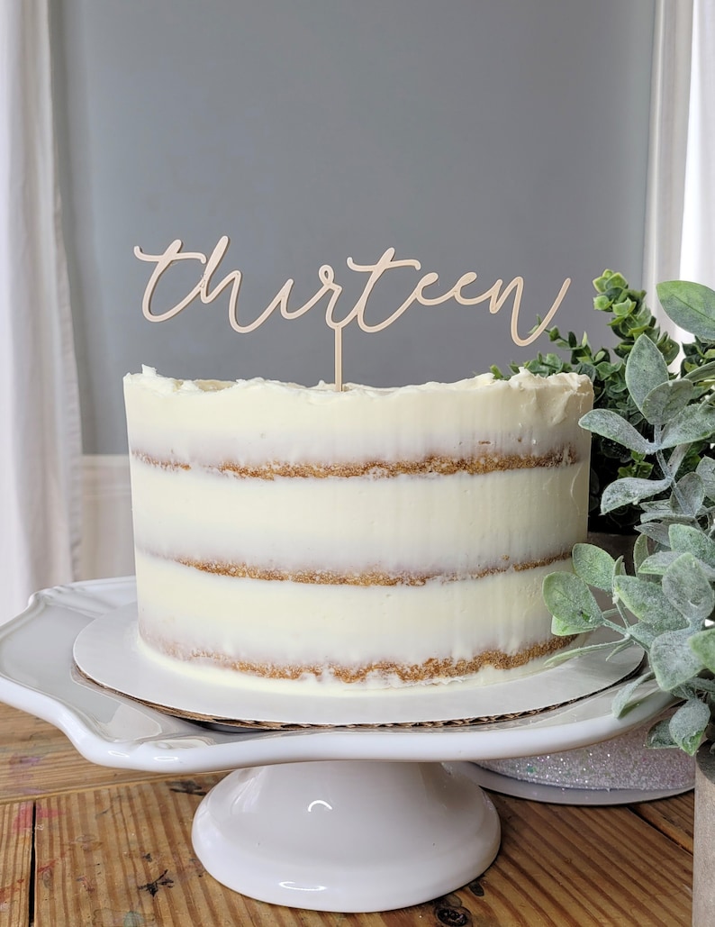 May include: A white cake with vanilla frosting and a wooden cake topper that says "thirteen".