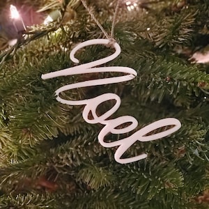 Puede incluir: Adorno blanco para árbol de Navidad con la palabra "Noel" escrita en letra cursiva.
