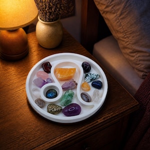 May include: A white circular tray shaped like a Leo zodiac symbol, containing an assortment of polished and raw gemstones in various colors including purple, green, blue, orange, and clear, arranged within the tray's curved compartments.