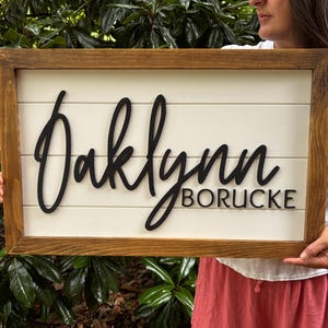 Op de afbeelding: Een rechthoekig houten bord met een donkerbruin gebeitste lijst en een witte achtergrond in shiplap-stijl. Het bord is voorzien van verhoogde zwarte letters met de naam Oaklynn in een groot cursief lettertype en BORUCKE in een schreefloos lettertype.