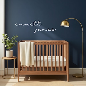 May include: A wooden cot with vertical slats, a white mattress, and a cream-coloured knit blanket. Above the cot, the name "emmett james" is written in white cursive script on a dark blue wall. A gold floor lamp and a small plant are nearby.