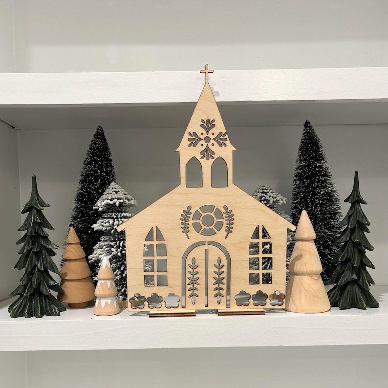 May include: Wooden cutout of a church with a snowflake design on the steeple. The church is surrounded by small wooden trees, some with snow on them.