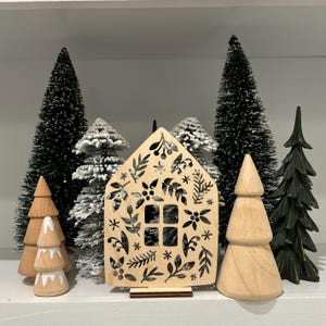 May include: A wooden cutout of a house with a black floral design. The house is surrounded by small artificial pine trees, some with white flocked tips.
