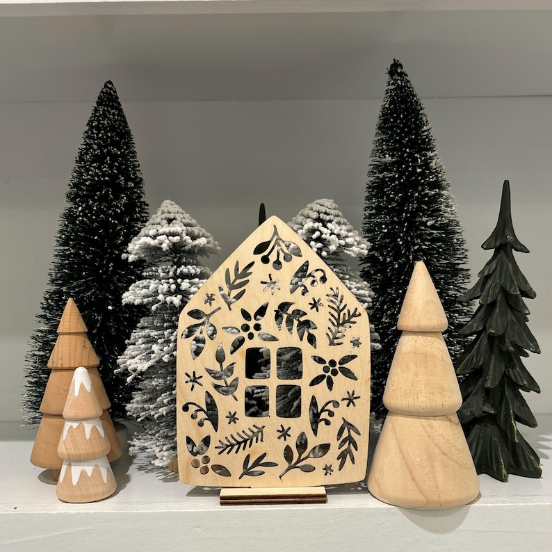 May include: A wooden cutout of a house with a black floral design. The house is surrounded by small artificial pine trees, some with white flocked tips.