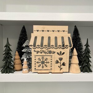 May include: A wooden cutout of a small shop with a snowflake and floral designs. The shop is on a shelf with small artificial pine trees.