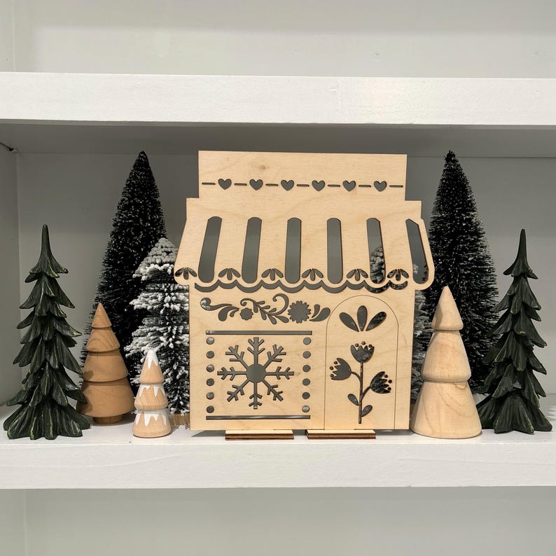 May include: A wooden cutout of a small shop with a snowflake and floral designs. The shop is on a shelf with small artificial pine trees.