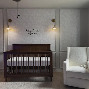 May include: A dark brown wooden crib with a mattress, set in a nursery. The crib is the focal point, with a white patterned mattress. The wall behind the crib has the names "Daphne Ryan" in cursive. A white armchair is to the right.
