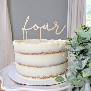 Può includere: Una torta bianca a tre piani con glassa alla vaniglia e un topper in legno che dice "quattro" in corsivo.